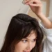 Close-up of a woman in a white waffle bathrobe applying a clear serum to her scalp with a glass dropper. A perfect visual for a guide on hair care routines, dry scalp treatments, or DIY hair growth oils.