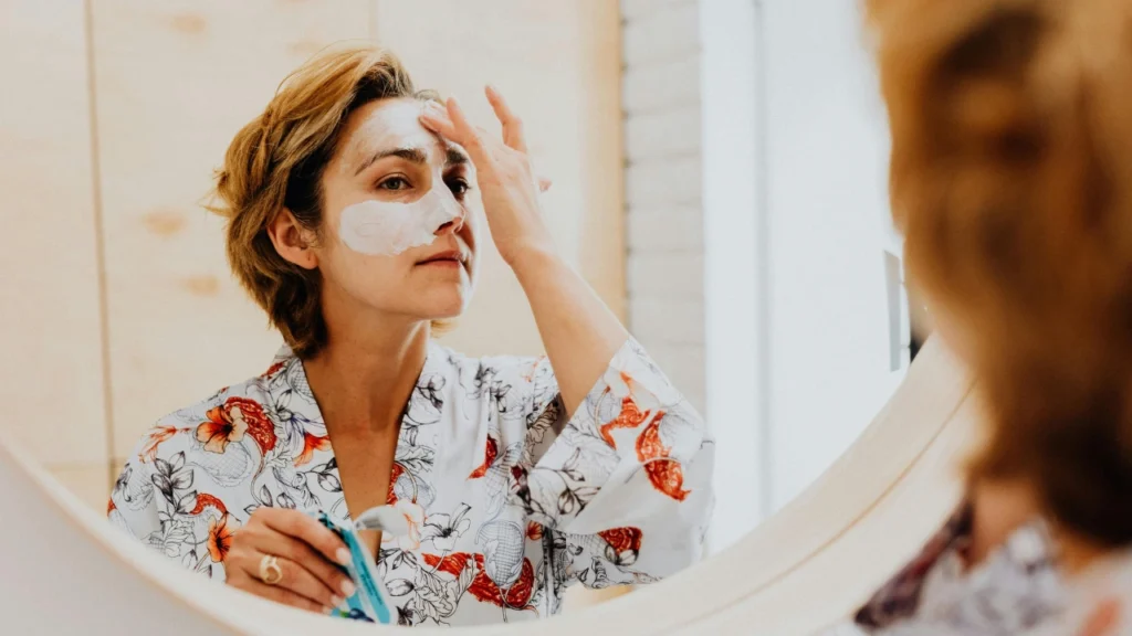 A person in a floral robe looking into a mirror while applying a white cream face mask to their forehead and nose.