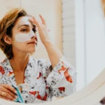A person in a floral robe looking into a mirror while applying a white cream face mask to their forehead and nose.