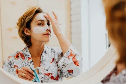 A person in a floral robe looking into a mirror while applying a white cream face mask to their forehead and nose.