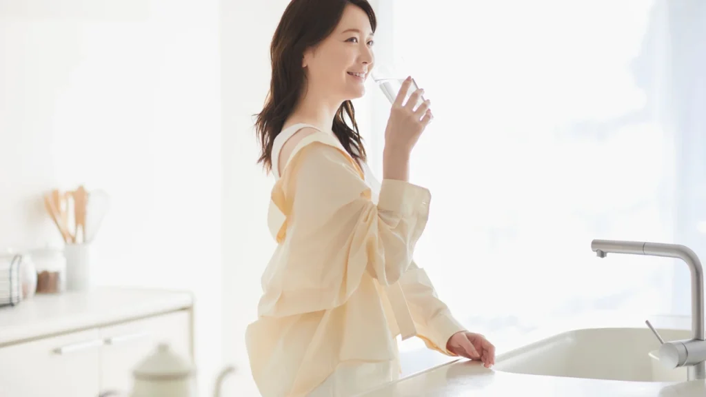 A smiling woman standing in a bright, modern kitchen takes a sip from a glass of water, highlighting hydration as a key healthy habit.