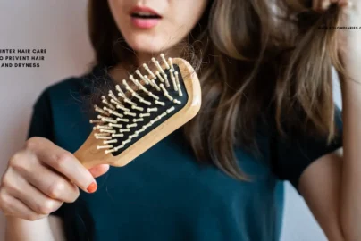This image shows a person looking concerned while holding a wooden hairbrush that has several strands of tangled hair caught in the bristles. The person has long brown hair and is wearing a dark green t-shirt. There is text on the left side of the image that reads: "BEST WINTER HAIR CARE TIPS TO PREVENT HAIR FALL AND DRYNESS". In the top right corner, the website "WWW.KGLOWDIARIES.COM" is displayed.