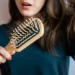 This image shows a person looking concerned while holding a wooden hairbrush that has several strands of tangled hair caught in the bristles. The person has long brown hair and is wearing a dark green t-shirt. There is text on the left side of the image that reads: "BEST WINTER HAIR CARE TIPS TO PREVENT HAIR FALL AND DRYNESS". In the top right corner, the website "WWW.KGLOWDIARIES.COM" is displayed.