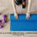 An overhead view of a person unrolling a blue yoga mat on a light wood floor, surrounded by workout gear including purple dumbbells and a pink water bottle, with text about how physical activity improves skin glow.