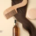 A top-down view of hair care essentials featuring a long strand of wavy brunette hair with a small braid, a wooden wide-tooth comb, an amber glass dropper bottle, and a round tan cosmetic jar on a neutral background.