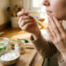 A close-up photograph of a woman applying a natural skincare treatment in her kitchen (`3094322938290813407.jpeg`). She wears a beige knit sweater and holds a ceramic spoon with dripping golden honey up to her chin, while her other hand gently touches her face. The wooden countertop in the blurred background features a jar labeled "RAW HONEY," a bowl of white yogurt, and a glass bowl filled with sliced cucumbers in water, creating a warm and rustic atmosphere.